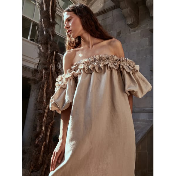A woman in a beige off-the-shoulder, ruffled linen dress stands outdoors by a tall palm tree, sunlight illuminating her face—evoking the warmth and elegance perfect for Fathers Day gifts 2026.