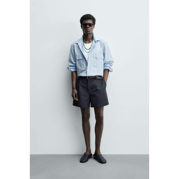 A man stands against a plain light grey wall wearing a light blue button-up shirt over a white vest, dark shorts, black loafers, sunglasses, and layered necklaces—a relaxed take on men's holiday fashion.