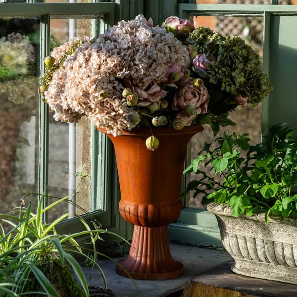 A brown vase filled with pink and green hydrangeas and peonies sits on a windowsill, bringing affordable interiors style next to a stone planter of leafy greens as sunlight streams through the window.