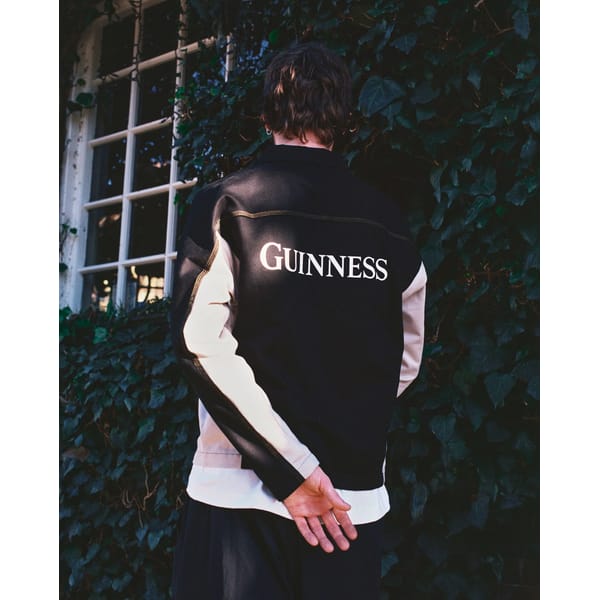 A person stands with their back to the camera, wearing a black and white football core jacket with GUINNESS written on the back, standing in front of a window and a wall covered in green ivy.