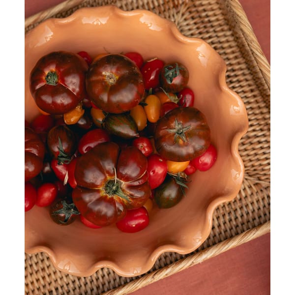 A peach-coloured, wavy-edged bowl filled with dark red heirloom tomatoes and assorted cherry tomatoes sits on a woven tray with a reddish table surface underneath—perfect for celebrating salad season.