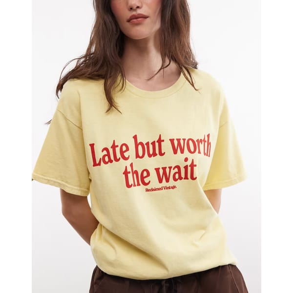 A woman wearing a yellow T-shirt with red text that reads Late but worth the wait, one of those cool slogan tops. Her arms are behind her back, and the background is plain white.