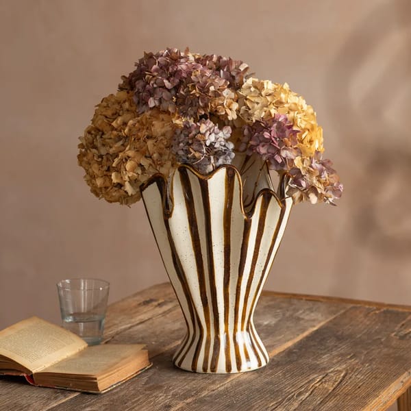 A striped ceramic statement vase with dried hydrangeas sits on a wooden table next to an open book and a glass of water, against a beige background.
