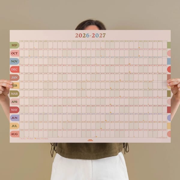 A person holding a large 2026-2027 wall calendar with months labelled on the left and days arranged in a grid, perfect for planning Summer Holidays With Children. The calendar features a light pastel colour scheme, while the person’s face is mostly hidden behind it.