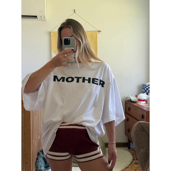 A person takes a mirror selfie wearing a loose white cool slogan T-shirt with the word MOTHER in bold black letters and red athletic shorts with white stripes, in a cosy, softly lit room.