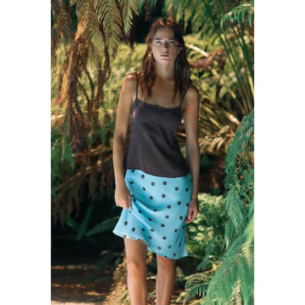 A woman stands among lush green ferns, wearing a black spaghetti-strap top and a light blue skirt with black polka dots—a perfect nod to turquoise fashion. Sunlight filters through the plants, illuminating her and the surroundings.