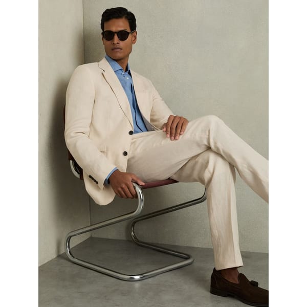 A man wearing sunglasses, a light beige suit, blue shirt, and brown loafers sits cross-legged on a modern chair against a neutral background, effortlessly capturing The Talented Mr Ripley style and exemplifying the essence of refined men’s summer dressing.