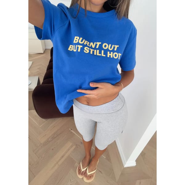 A woman stands indoors wearing one of those cool slogan tees—a blue T-shirt that says BURNT OUT BUT STILL HOT—paired with grey shorts and beige flip-flops. She is holding her shirt up slightly, revealing part of her midriff.