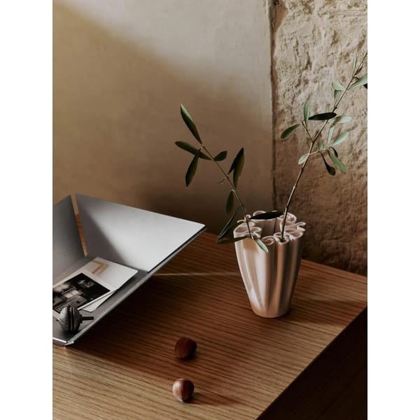 A small ceramic statement vase with green branches sits on a wooden table next to a tray holding papers and a photo; two small brown objects rest on the table. A textured wall is in the background.