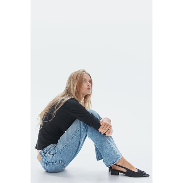 A person with long blonde hair, wearing a black long-sleeved top, blue jeans, and black ballet flats—styled in the effortless elegance of minimalist high street brands—sits on the floor with knees drawn up and arms wrapped around them, looking slightly upwards.