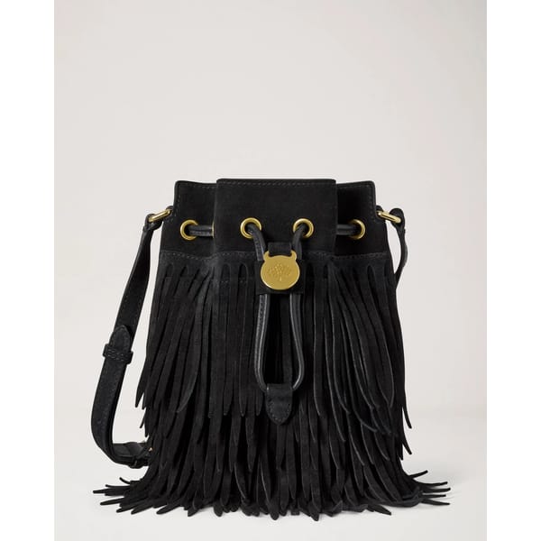 Black suede bucket bag with long fringe, gold eyelets, a gold medallion, and an adjustable shoulder strap—this fringed bag is the perfect statement accessory for cool girls to carry into autumn, displayed against a plain light background.