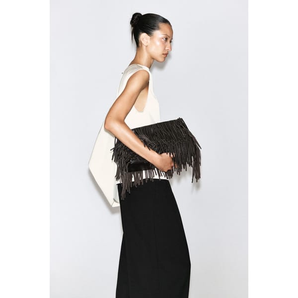 A woman with tied-back hair wears a sleeveless white top and wide black trousers, holding a large, black fringed clutch bag—a perfect accessory for cool girls—against a plain background.