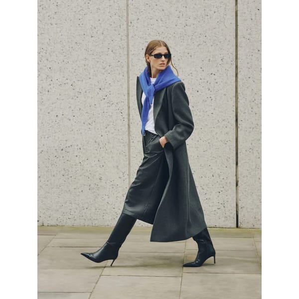 A woman in a long dark coat, black heeled boots, and sunglasses walks outside. She has an International Klein Blue jumper draped over her shoulders and wears cropped black trousers and a white top. The background is a light stone wall.