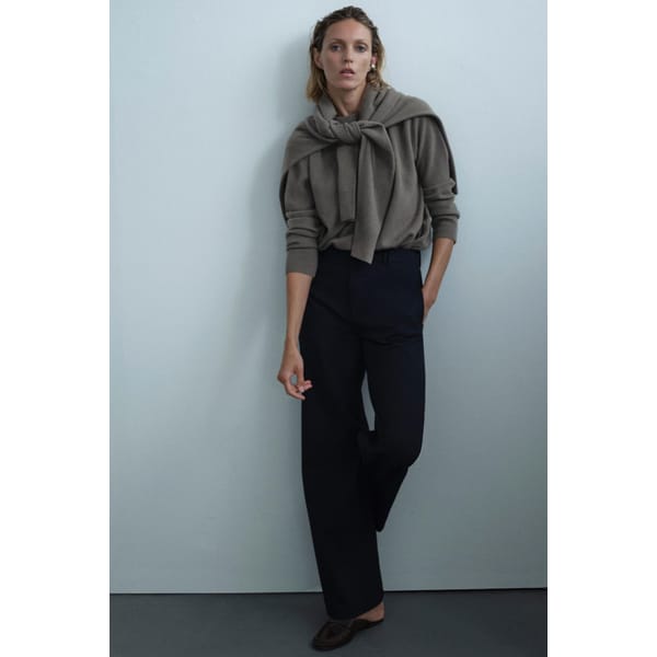 A woman stands against a plain wall wearing a taupe jumper from one of her favourite minimalist high street brands, with another jumper draped over her shoulders, black wide-leg trousers, and brown shoes. She looks relaxed with one hand in her pocket.