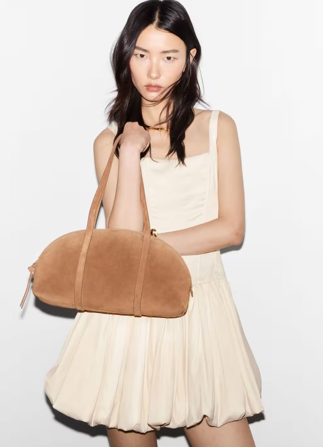 model wearing cream dress and holding tan bowling bag