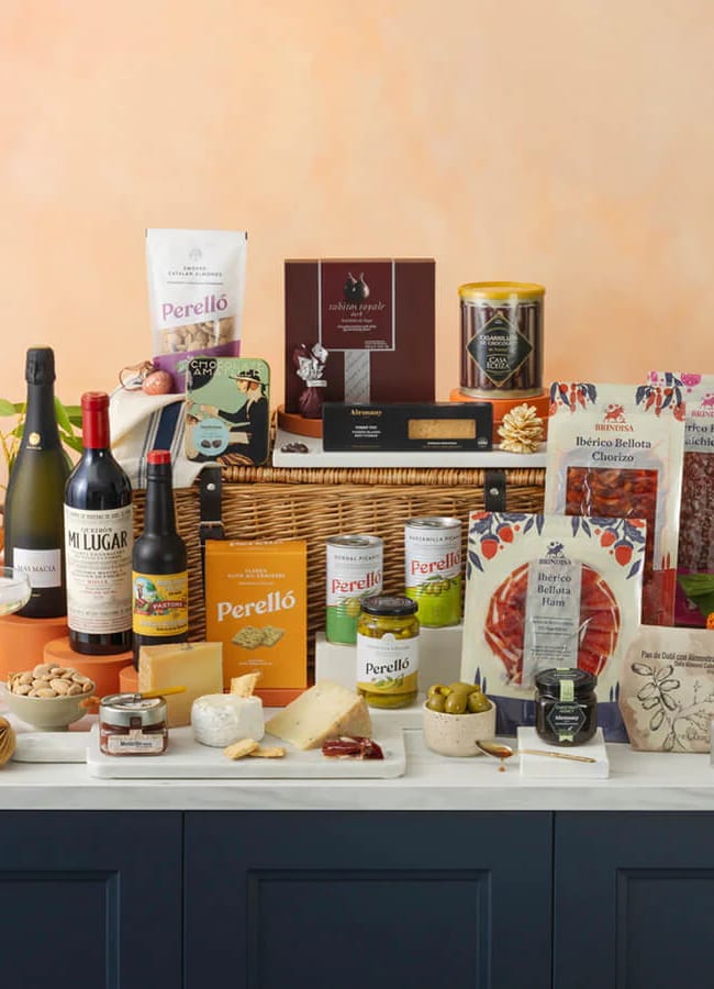 A luxurious Christmas hamper with assorted gourmet foods, wine, cheese, olives, chocolates, crackers, nuts, and charcuterie arranged neatly on a counter with colourful flowers, candles, and a wicker basket in the background.