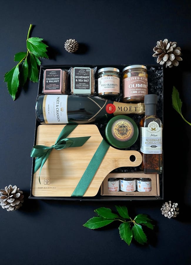 A luxurious Christmas hamper featuring a bottle of Moët champagne, gourmet snacks, jars of condiments, a cheese board tied with a green ribbon, plus festive pine cones and leaves—all artfully arranged on a black background.
