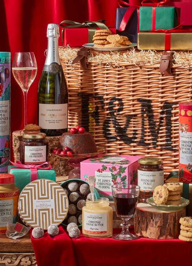 A festive Christmas hampers selection filled with assorted gourmet treats—champagne, wine, biscuits, preserves, pudding, and chocolates—displayed with glasses and wrapped presents against a vibrant red backdrop.
