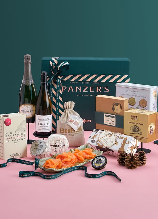 A festive Christmas hamper with wine, smoked salmon, biscuits, chocolates, panettone, sauces, and crackers is arranged on a pink surface with a green background. Four tall, colourful candles and pinecones decorate the scene.