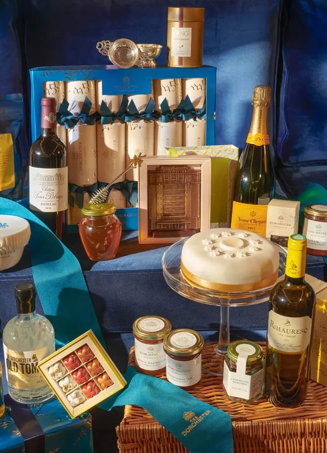 A luxurious Christmas hamper featuring assorted food and drink items—wine, champagne, spirits, chocolates, preserves, honey, biscuits, and a decorated cake—all beautifully arranged on a wicker basket with blue ribbon atop a blue velvet background.