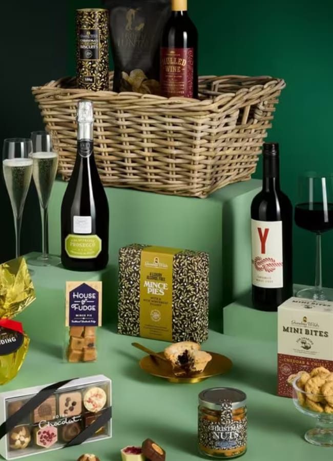A luxurious Christmas hamper featuring wine, champagne, glasses, mince pies, pudding, biscuits, fudge, chocolate, nuts, and more treats arranged on green platforms against a dark green background.