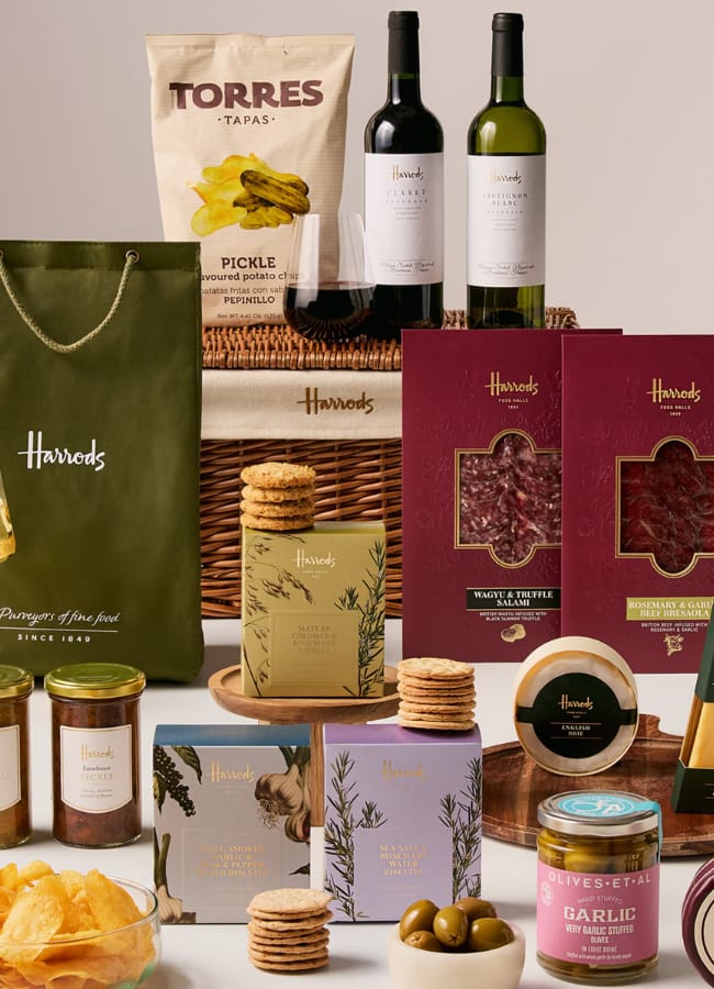 A luxury Harrods Christmas hamper with wine bottles, crisps, biscuits, jars of spreads, olives, cheese, charcuterie, and chocolates in elegant packaging displayed on and around a wicker basket.