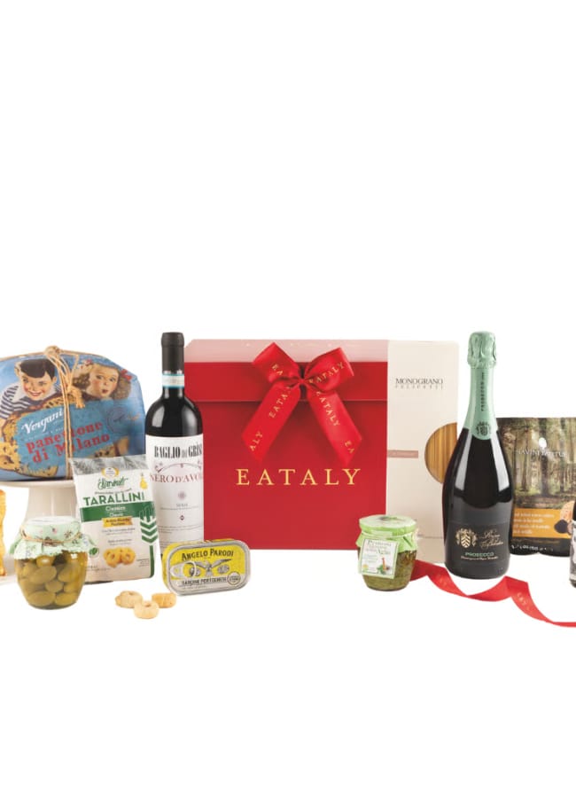 A festive Eataly Christmas hampers gift box with a red ribbon is surrounded by Italian treats: panettone, wine, sparkling wine, pasta, olives, tarallini, honey, sauce, and spreads—all displayed on a white background.