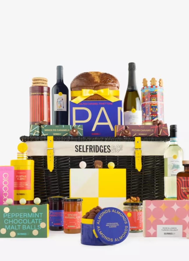A luxury Selfridges Christmas hamper filled with assorted gourmet items including wine, chocolates, tea, biscuits, preserves, and festive treats—all arranged neatly in front of a black wicker basket with yellow accents.