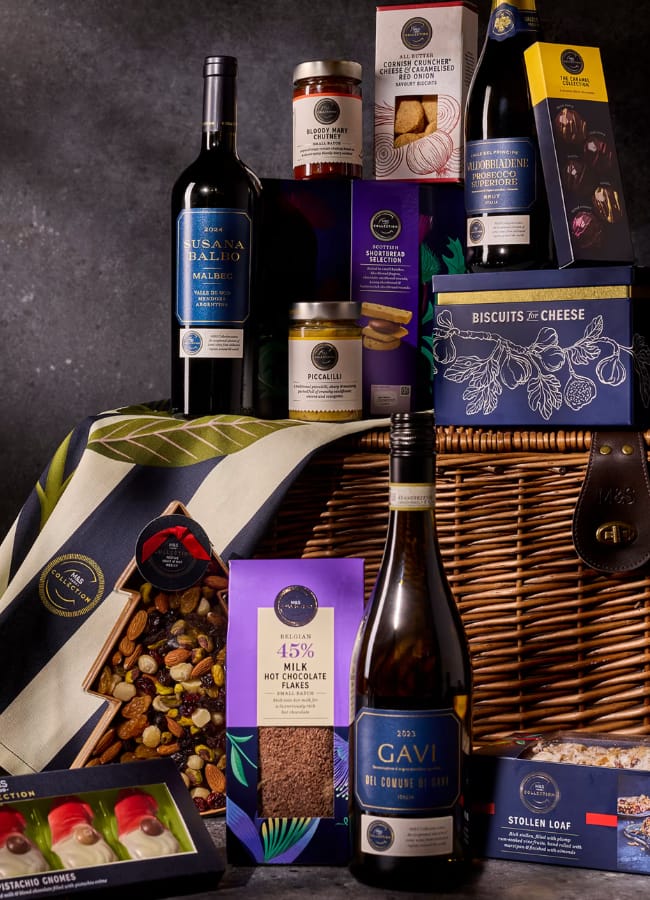 A luxury Christmas hamper includes red and white wine, biscuits, preserves, crackers, chocolates, stollen loaf, fruit and nut mix, cheeses, jars, and a decorative cloth, all beautifully arranged beside a wicker basket.