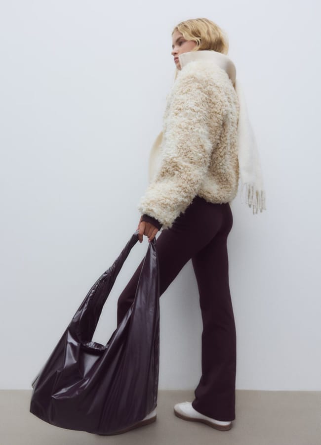 A person in a fluffy cream jacket, black trousers from the latest activewear collection, and white trainers stands against a plain wall, holding a large black tote bag and wearing a white scarf.