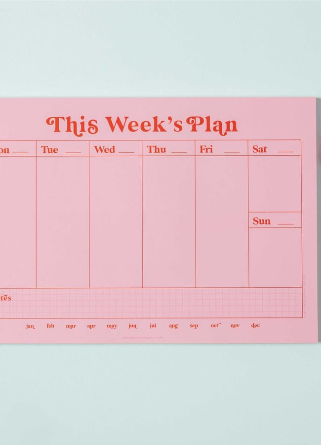 A pink weekly planner inspired by 2026 diaries, featuring columns for each day, a notes section at the bottom, an orange pen, and a pink hexagonal rubber on a light blue surface.