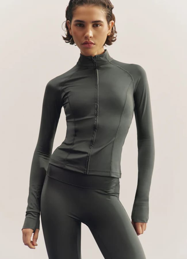 A person with short hair wears a dark, fitted, full-zip sports jacket and matching leggings—showcasing the latest activewear—whilst standing against a light background.
