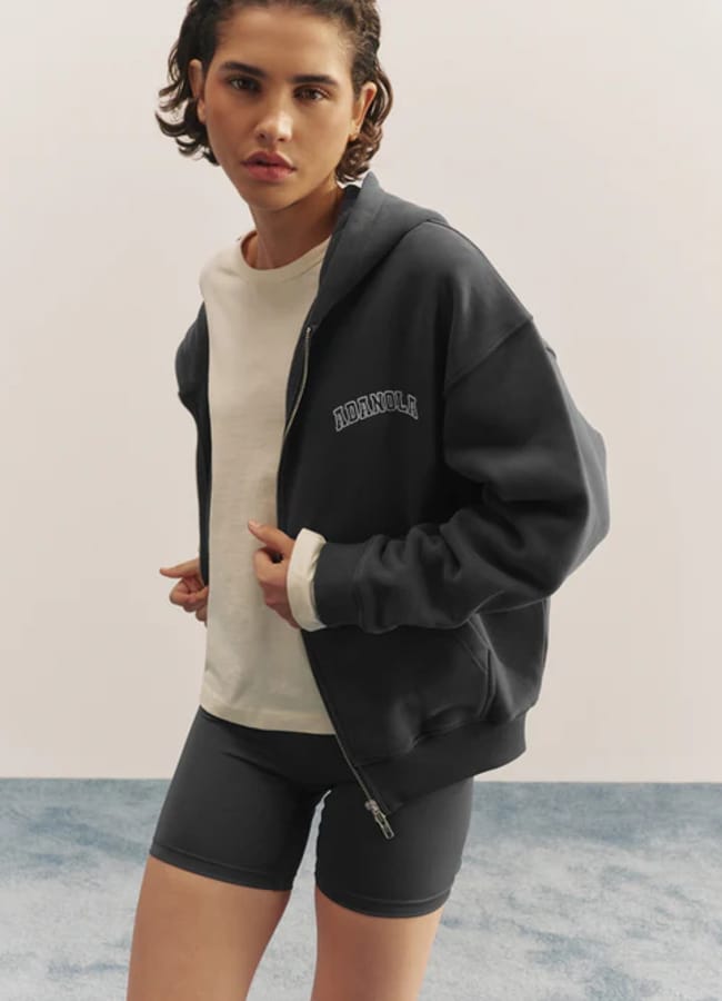 A person with short, wavy dark hair wears the latest sportswear: a white T-shirt, dark zip-up hoodie, and matching cycling shorts. They stand indoors on a blue carpet against a plain light background.