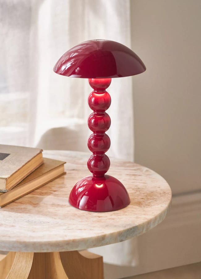 A modern red table lamp, inspired by the Unexpected Red Theory, features a dome-shaped shade and stacked sphere base on a marble side table beside two closed books—an eye-catching 2026 interior design accent set against a white curtain backdrop.