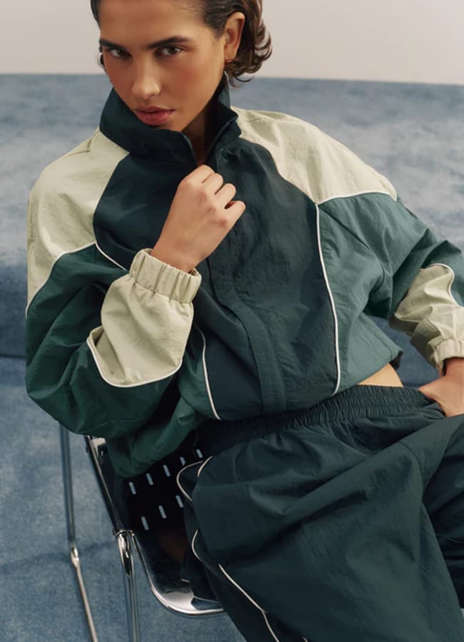 A person wearing the latest activewear—a green and beige colour-blocked tracksuit—sits confidently on a black chair, looking at the camera in a room with a blue carpet.