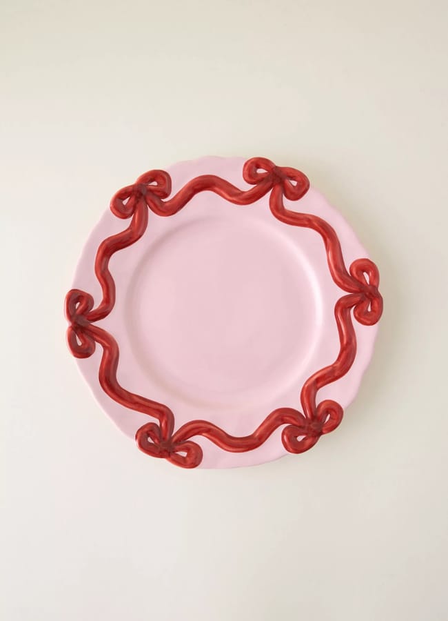 A round, light pink ceramic plate with a raised, wavy red ribbon design forming bows around the edge—perfect for adding charm to your Christmas table setting—set against a plain off-white background.