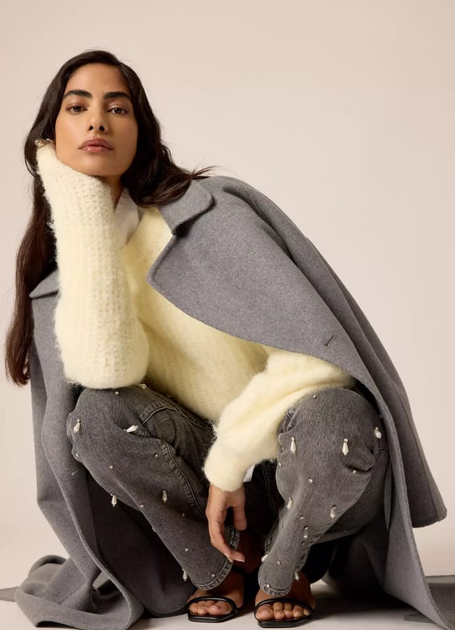 A woman poses squatting, wearing a soft cream jumper, embellished jeans, a grey coat draped over her shoulders, and black sandals, resting her head on her hand against a plain background.