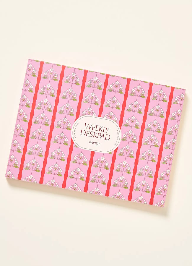 A rectangular weekly deskpad with a pink cover featuring a repeating floral pattern and a white oval label in the centre that reads WEEKLY DESKPAD PAPER—perfect to complement your 2026 diaries.