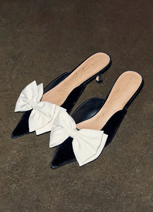 A pair of black pointed-toe heeled mules with large white bows on the front, displayed on a dark textured surface—perfect for elevating your bow fashion style.