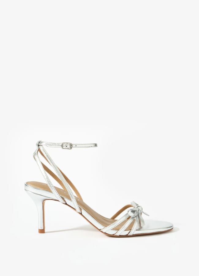 A silver high-heeled sandal with an ankle strap, thin criss-crossing straps, an open toe, and a small bow detail on the front—perfect for bow fashion lovers. The stiletto heel and straps feature a shiny metallic finish.