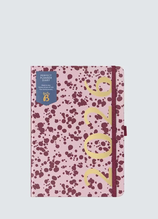 A pink 2026 diary with dark maroon splatter patterns, a zip fastening on the right, and large yellow numbers 2026 printed diagonally on the cover. Perfect for fans of stylish 2026 diaries, it features a blue label on the top left corner.