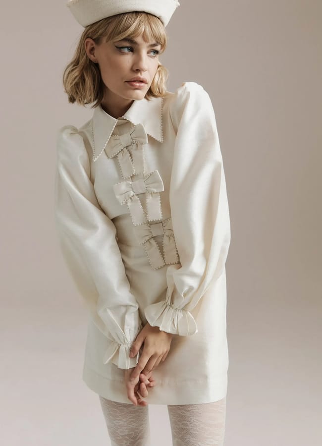 A woman wearing a vintage-inspired, long-sleeved ivory mini dress with bow fashion details and pearl accents stands in a neutral studio. She has a beige pillbox hat, blonde hair with fringe, and lace-patterned white tights.