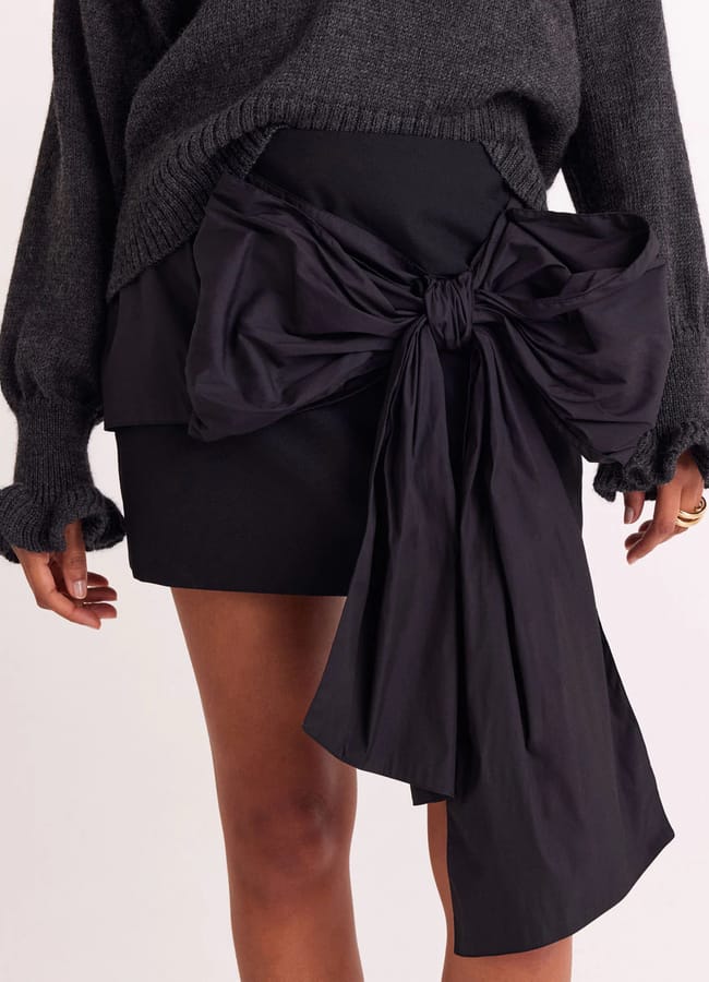 A person wearing a dark jumper and a black mini skirt featuring a large, dramatic bow with long, flowing tails on the front—a striking example of bow fashion. Only the lower half of the body is visible.
