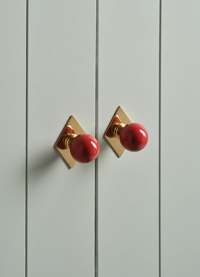 Two red round door knobs with gold diamond-shaped backplates add a touch of Unexpected Red Theory to this light grey panelled cabinet, making it a chic statement for modern interior design.