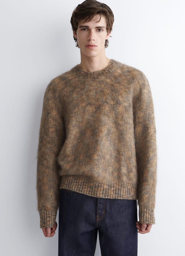 A young person with short brown hair is standing against a white wall, wearing a fuzzy brown and beige men's jumper with long sleeves and dark blue jeans. They have a neutral expression and face the camera directly.