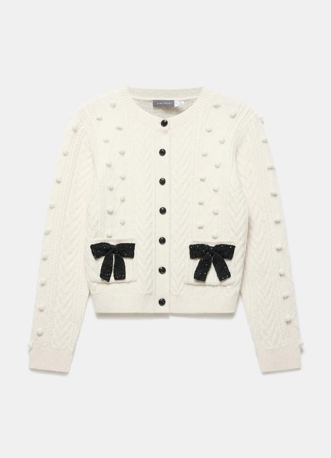 A cream-coloured, long-sleeve knitted cardigan with a textured bobble pattern, black buttons down the front, and two front pockets each decorated with a black fabric bow—a charming nod to bow fashion.