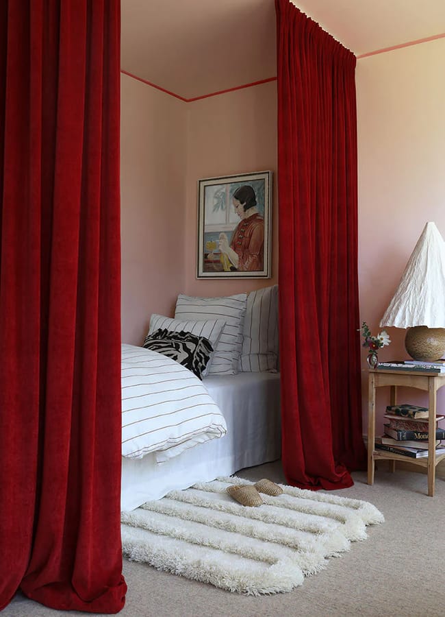 A cosy bedroom inspired by Unexpected Red Theory, featuring bold red curtains framing a bed with white and black striped bedding, a fluffy rug, a sculptural lamp on the bedside table, and a portrait adorning the pale pink wall—a 2026 interior design vision.