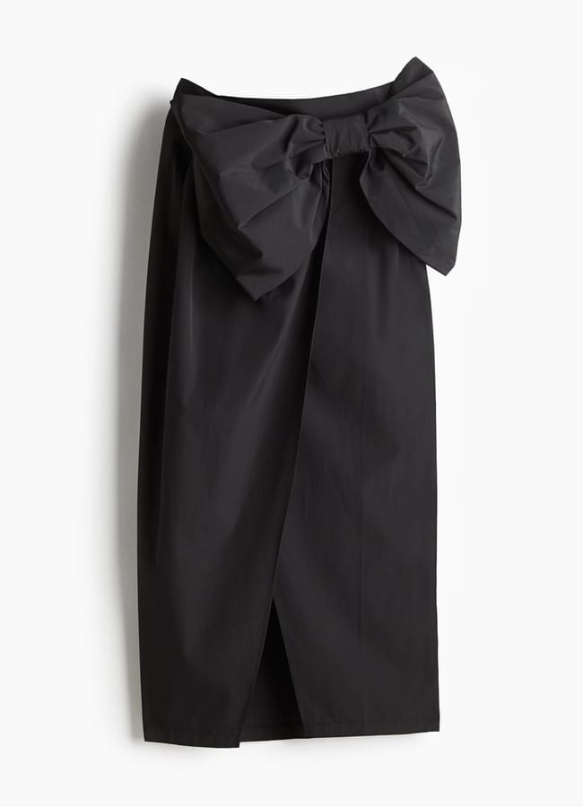 A black knee-length skirt featuring a large, structured bow at the waist showcases iconic bow fashion. With a subtle front overlap slit and smooth, slightly shiny fabric, this elegant skirt is displayed against a white background.