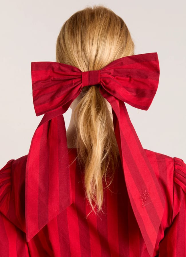 A person with long blonde hair tied in a low ponytail, adorned with a large red striped bow—a striking touch of bow fashion. The matching red striped garment stands out against the plain light grey background.