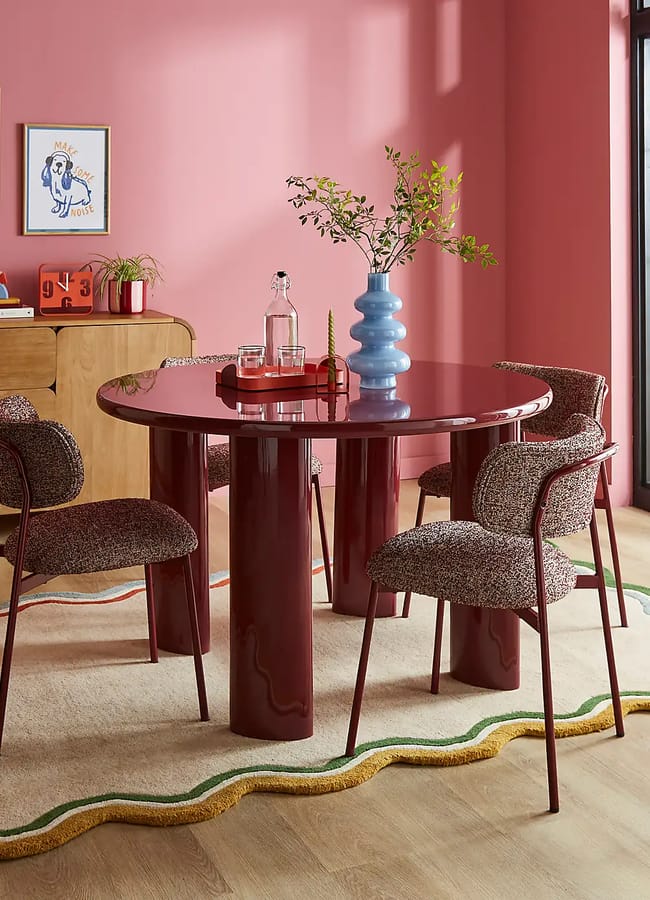 A modern dining area showcasing Unexpected Red Theory with a glossy burgundy table, four textured chairs, pink walls, framed artwork, a wooden sideboard, and large windows offering a garden view—perfect inspiration for 2026 interior design.